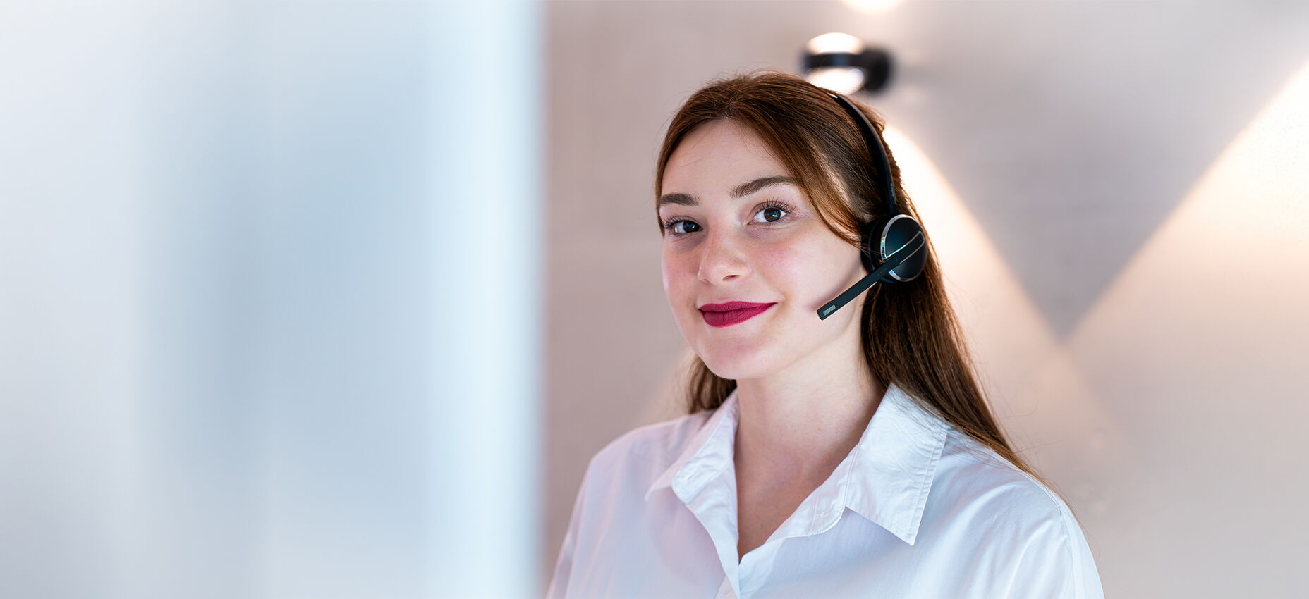 Junge Frau mit Headset und weißer Bluse blickt freundlich in die Kamera, vor neutralem Hintergrund.“