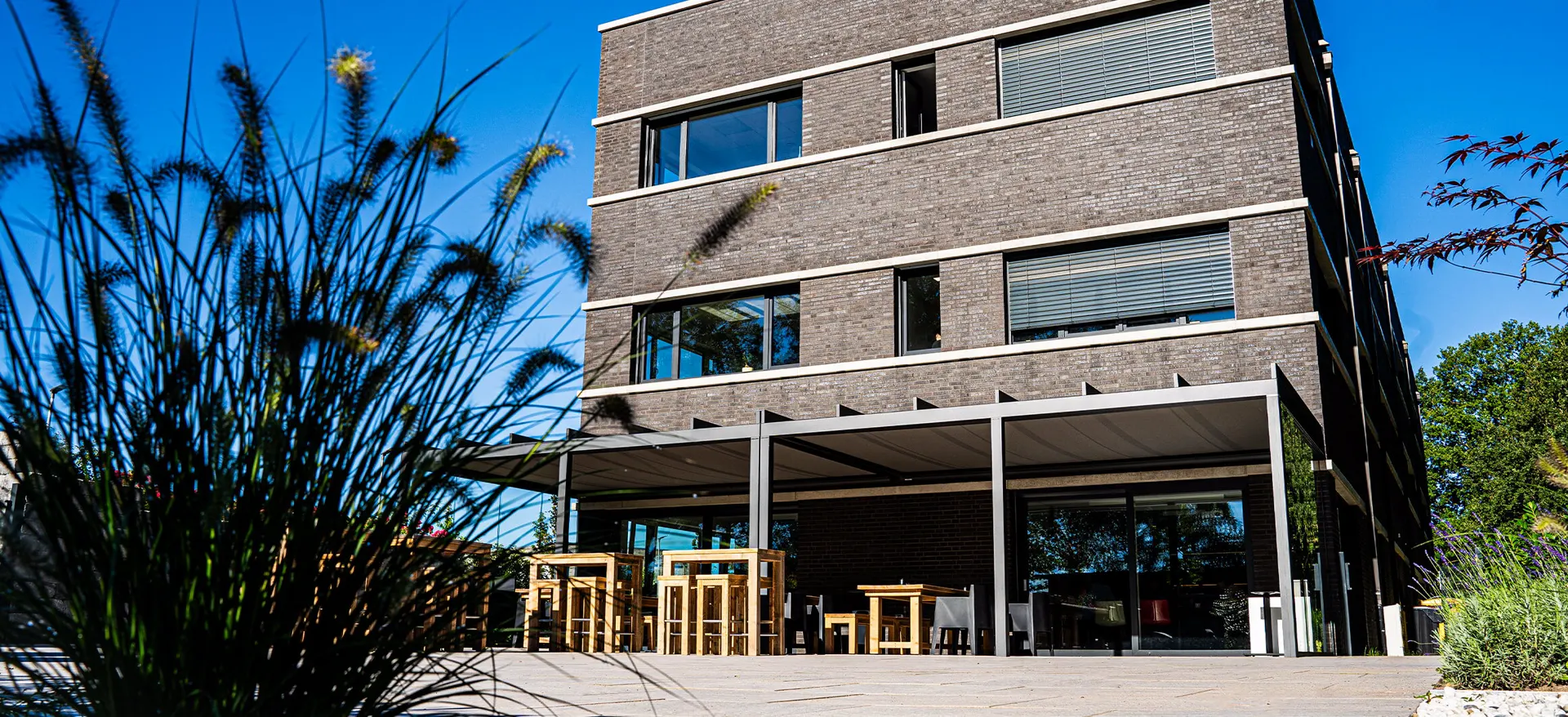 Moderne Gebäuderückseite mit überdachter Terrasse und Holzsitzgruppen, umgeben von Pflanzen. Das Bild zeigt die Rückseite eines modernen Bürogebäudes aus dunklem Klinker mit großen Fenstern und Jalousien.
