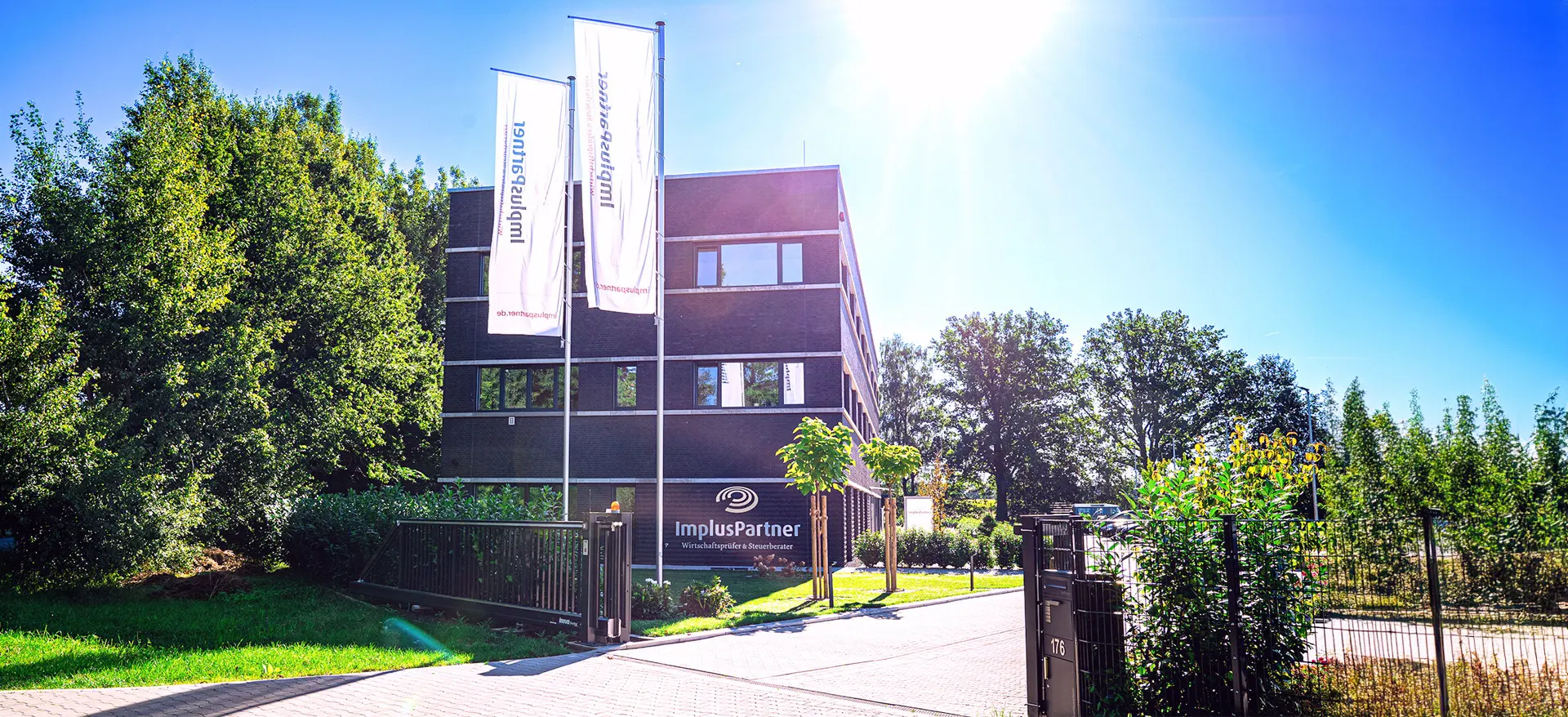 Firmensitz von ImplusPartner mit dunkel verklinkertem Gebäude, Fahnenmasten und gepflegtem Außengelände