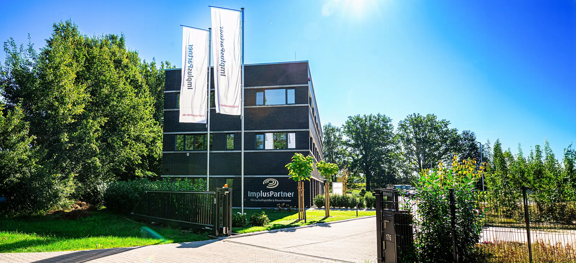 Firmensitz von ImplusPartner mit dunkel verklinkertem Gebäude, Fahnenmasten und gepflegtem Außengelände