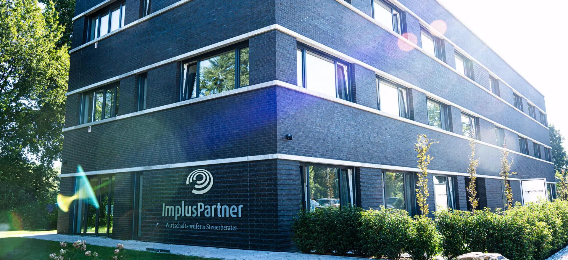 Firmengebäude von ImplusPartner mit Logo an der Fassade und Garten im Vordergrund.“